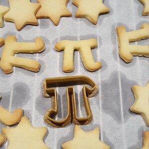 Peut inclure: Un emporte-pièce en forme de symbole mathématique pi de couleur dorée, entouré de biscuits cuits en forme d'étoiles et de la lettre pi.