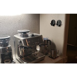 May include: A stainless steel espresso machine and coffee grinder sit on a countertop. A milk frother and a metal mug are also visible. Two black hooks are mounted on the wall. The scene is illuminated by a soft overhead light.