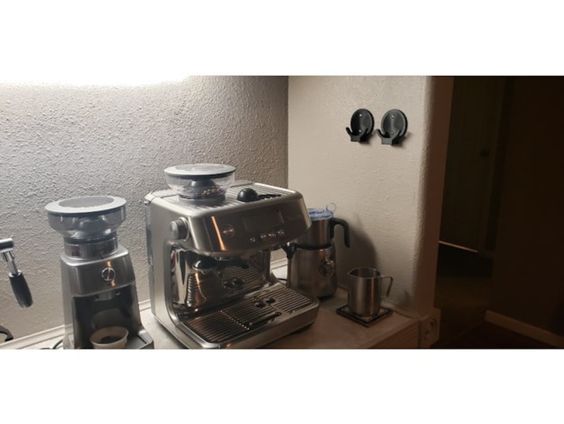 May include: A stainless steel espresso machine and coffee grinder sit on a countertop. A milk frother and a metal mug are also visible. Two black hooks are mounted on the wall. The scene is illuminated by a soft overhead light.