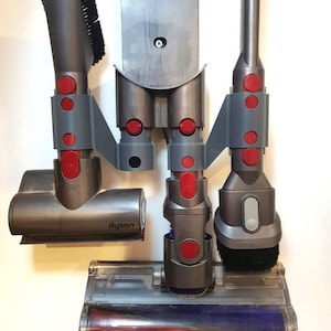 Pode incluir: Um suporte de parede cinza com um logotipo Dyson e quatro acessórios cinzentos e vermelhos para um aspirador de pó. Os acessórios incluem uma ferramenta para fendas, uma escova para pó, uma ferramenta combinada e uma cabeça de escova motorizada.