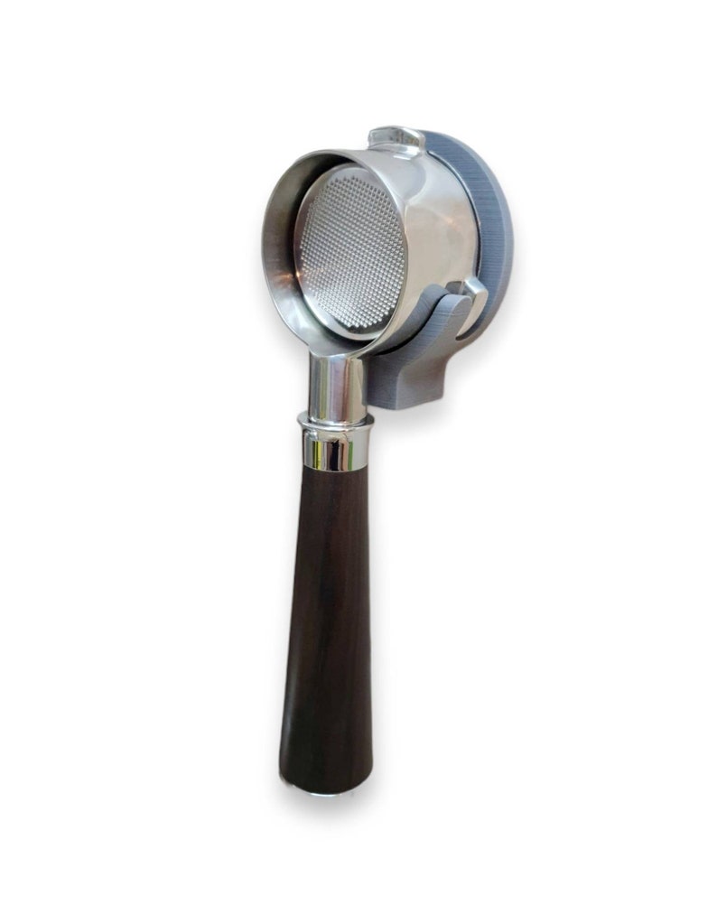 May include: A stainless steel portafilter with a dark brown wooden handle. The portafilter is held in a gray holder. The filter basket is visible inside the portafilter. The handle is a dark brown color.