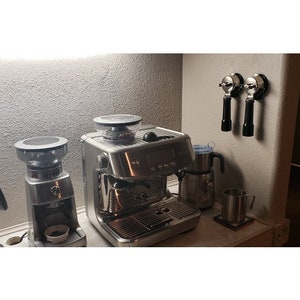 May include: A stainless steel espresso machine, coffee grinder, and milk frother set on a countertop. Two portafilters hang on the wall. A metal mug sits on a coaster. The appliances have black and silver accents.