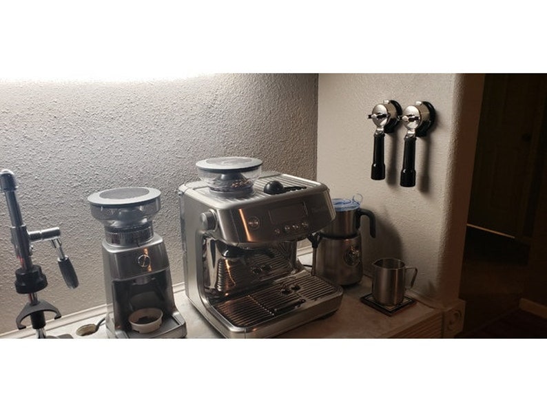 May include: A stainless steel espresso machine, coffee grinder, and milk frother set on a countertop. Two portafilters hang on the wall. A metal mug sits on a coaster. The appliances have black and silver accents.