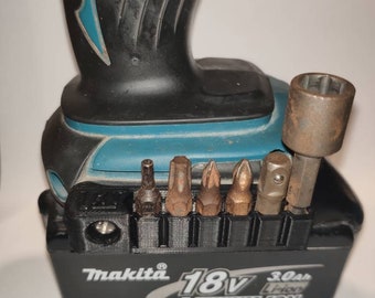 May include: A black and teal Makita 18V 3.0Ah Lithium-Ion battery with a 3D printed black holder for various screwdriver bits. The holder has 8 slots for bits, and there are 7 bits in the slots. The bits are silver, gold, and black.