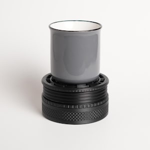 May include: A gray ceramic mug with a white interior and a thin black rim, resting on top of a black cylindrical herb grinder featuring a textured, knurled grip pattern around its base and a slotted top surface.