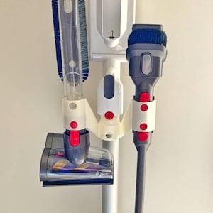 Peut inclure: Un support mural blanc pour accessoires d'aspirateur avec trois accessoires. Le support a une base blanche et un poteau argenté. Les accessoires comprennent un embout plat, une brosse à poussière et un embout combiné. Les outils sont tous noirs avec des boutons rouges.