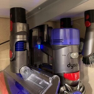 May include: A set of three Dyson vacuum cleaner attachments mounted on a wall. The attachments include a crevice tool, a dusting brush, and a motorized brush head. The attachments are all gray and purple with red accents.
