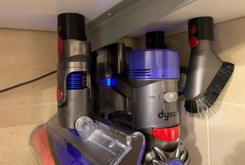 May include: A set of three Dyson vacuum cleaner attachments mounted on a wall. The attachments include a crevice tool, a dusting brush, and a motorized brush head. The attachments are all gray and purple with red accents.