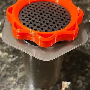 Può includere: Un portafiltro per macchina da caffè arancione e nero. La parte arancione ha un design circolare a coste con una serie di fori al centro. Il portafiltro è posizionato sopra un contenitore grigio scuro.