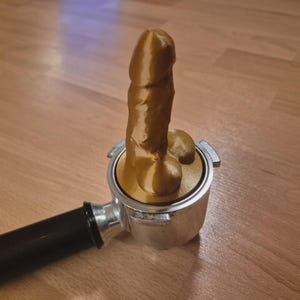 May include: A novelty item featuring a gold-colored, phallic-shaped object inserted into a silver espresso machine portafilter. The handle of the portafilter is black, and the item rests on a wooden surface.