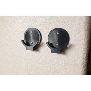 May include: Two black, wall-mounted hooks. Each hook has a circular base with a curved hook extending downward. The hooks are attached to the wall with screws. The hooks are made of a matte black material.