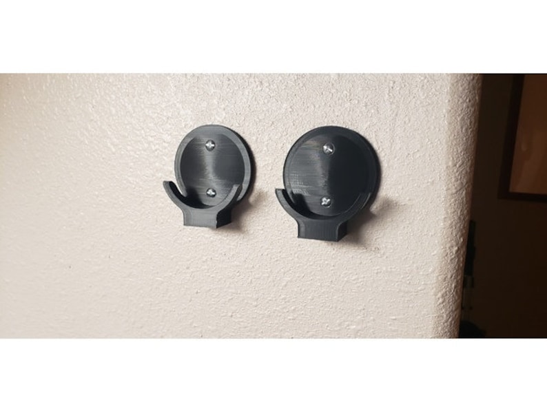May include: Two black, wall-mounted hooks. Each hook has a circular base with a curved hook extending downward. The hooks are attached to the wall with screws. The hooks are made of a matte black material.