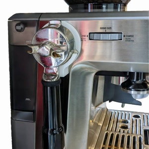 May include: Stainless steel espresso machine with a black handle and a clear bean hopper. The machine has a grind size dial and a portafilter attached. The hopper is filled with coffee beans.