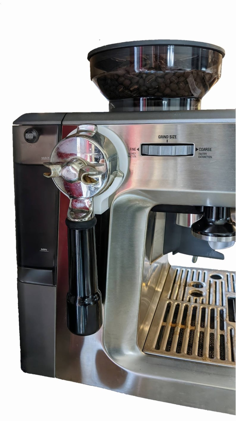 May include: Stainless steel espresso machine with a black handle and a clear bean hopper. The machine has a grind size dial and a portafilter attached. The hopper is filled with coffee beans.