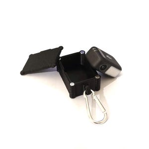 May include: A black rectangular case with a hinged lid and a silver carabiner clip. A small black camera with a white light sits beside the open case. The case is open, showing its interior.