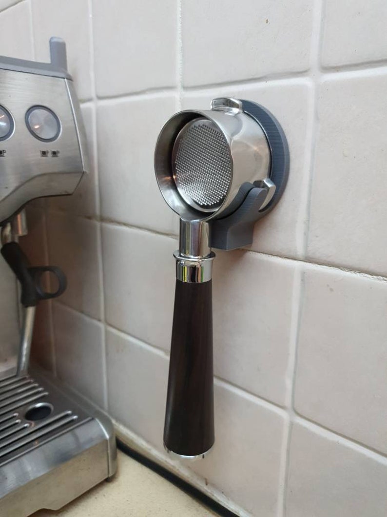 May include: A stainless steel portafilter with a dark brown handle is mounted on a gray holder attached to a tiled wall. The portafilter is designed for use with an espresso machine, visible on the left.