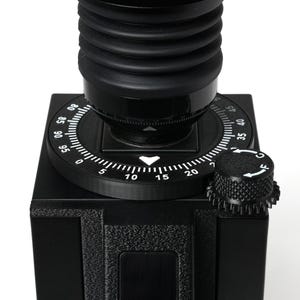 May include: A black camera bellows attachment featuring a circular dial with white numerical markings from 0 to 95. A smaller, textured black adjustment knob with white C and F markings sits on the side of the square base unit.