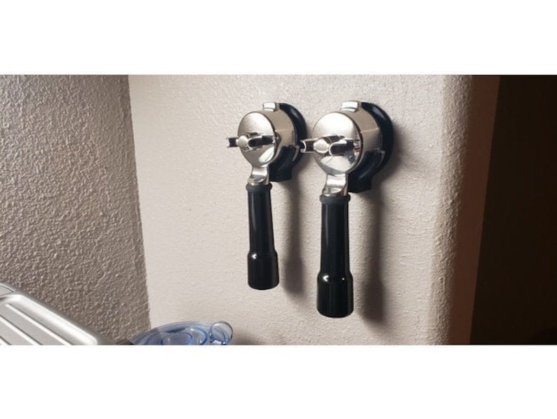 May include: Two espresso machine portafilters mounted on a light-colored wall. The portafilters have a shiny, metallic finish with black handles. The image is well-lit, highlighting the details of the coffee-making equipment.