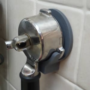 May include: A stainless steel espresso machine portafilter holder mounted on a gray holder. The portafilter has a black handle and is attached to a gray wall mount. The holder is attached to a white tiled wall.