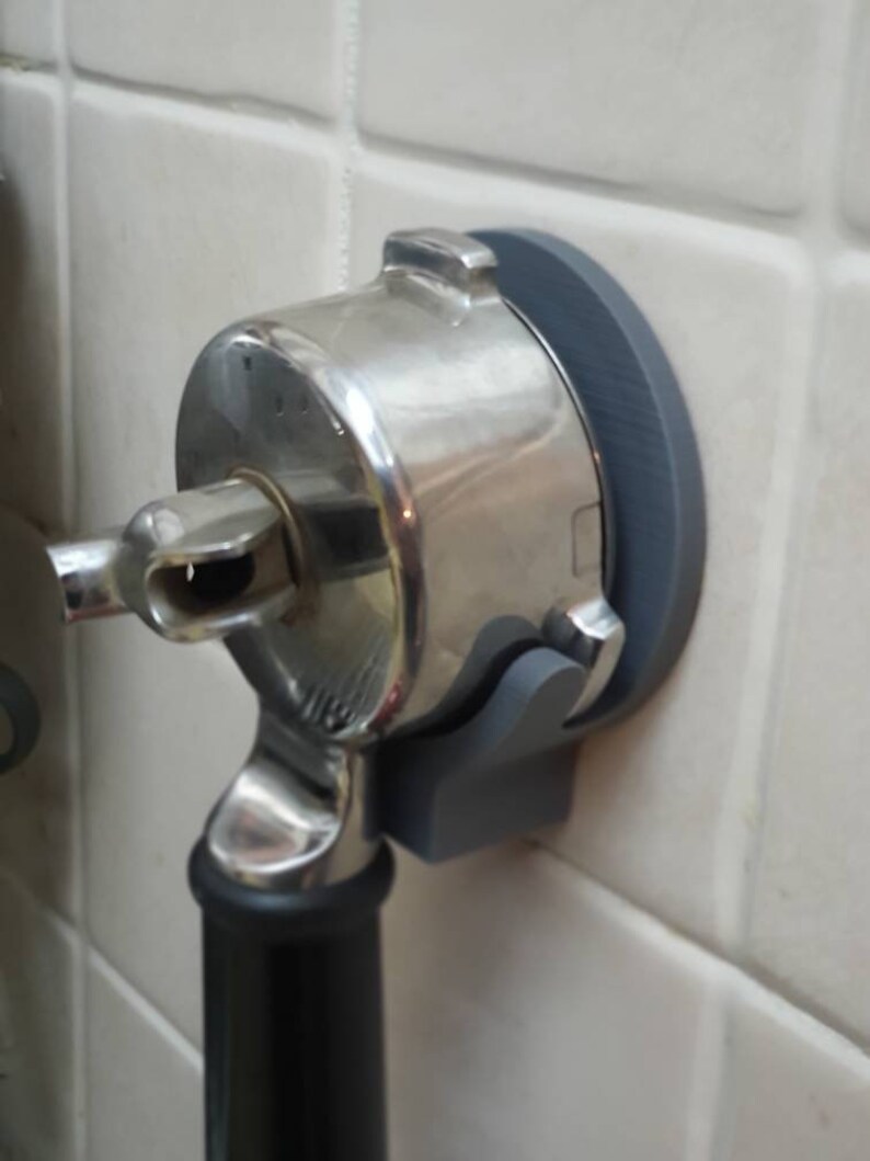 May include: A stainless steel espresso machine portafilter holder mounted on a gray holder. The portafilter has a black handle and is attached to a gray wall mount. The holder is attached to a white tiled wall.