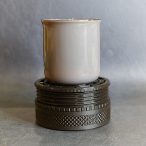 May include: A light grey cylindrical candle vessel with a smooth finish sits atop a dark brown circular base. The base features a textured, grid-like pattern around its side and a series of horizontal ridges, providing a sturdy foundation.