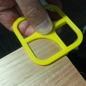 May include: A bright yellow plastic tool with four square openings. The tool is being held by a hand and is resting on a wooden surface.
