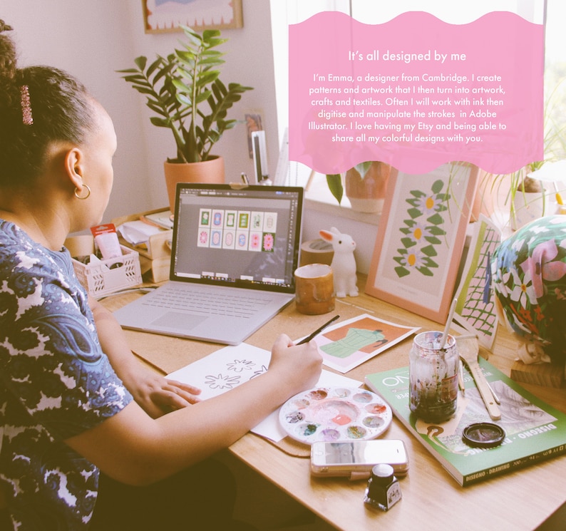 May include: A woman sits at a desk working on a colorful floral design. She is using a paintbrush and a palette of watercolors. There are other art supplies on the desk, including a laptop, a mug, a framed print, and a book. The text "It's all designed by me" is visible in the image.