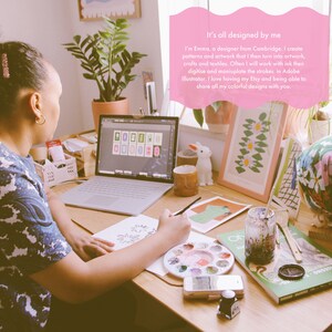 Op de afbeelding: Een vrouw zit aan een bureau en werkt aan een tekening met een penseel en een aquarelpalette. Ze heeft een laptop open voor zich, waarop een raster met kleurrijke ontwerpen te zien is. Achter haar hangen verschillende andere kleurrijke ontwerpen ingelijst aan de muur. De tekst "It's all designed by me" is in roze op een roze achtergrond geschreven.