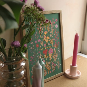 May include: A framed print with a green background and gold floral illustrations. The print says "Beauty & Power" in gold lettering. Two striped candles are in front of the print, and a pink candle is in a holder on a cork surface.