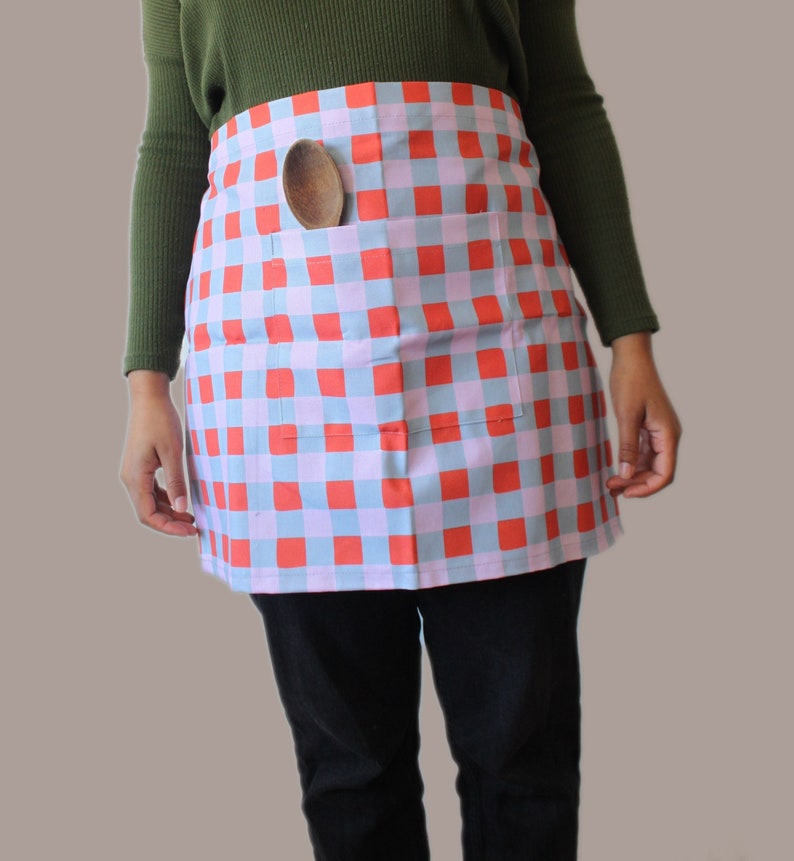 A red, blue and lilac half apron. The apron is made from a cotton linen blend and there is a pocket on the front. Inside the pocket is a wooden spoon which fits snuggly inside.