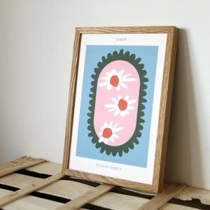 May include: A framed print with a blue background and a pink oval shape with three white flowers with red centers. The oval is surrounded by a dark green scalloped border. The text "LESLIE" and "FLOWER MARKET" are printed in black at the top and bottom of the print.