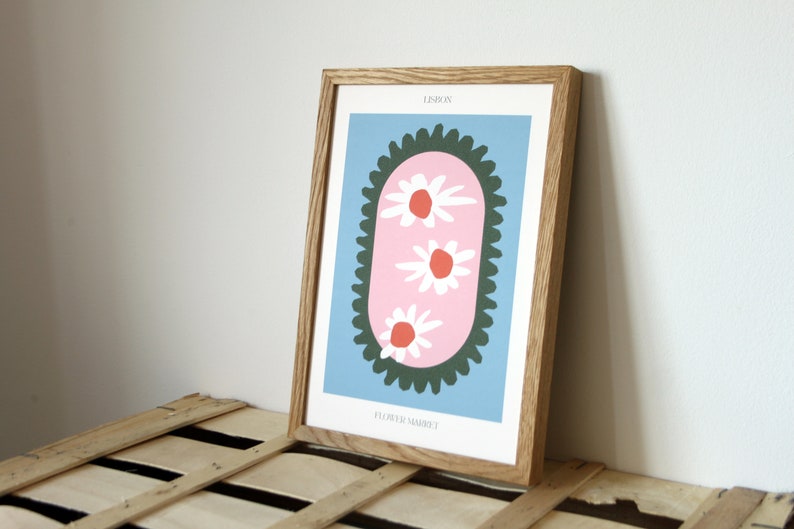 May include: A framed print with a blue background and a pink oval shape with three white flowers with red centers. The oval is surrounded by a dark green scalloped border. The text "LESLIE" and "FLOWER MARKET" are printed in black at the top and bottom of the print.