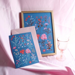 Op de afbeelding: Een blauwe kaart met een bloemenpatroon en de tekst "Happy Birthday" in rood. De kaart zit in een roze envelop en ligt naast een ingelijst afdruk met een vergelijkbaar bloemenpatroon en de tekst "Wee Was Born".