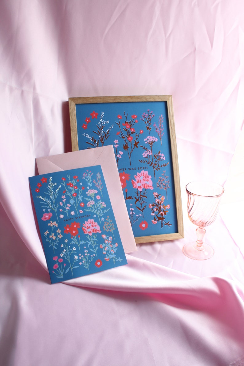 Op de afbeelding: Een blauwe kaart met een bloemenpatroon en de tekst "Happy Birthday" in rood. De kaart zit in een roze envelop en ligt naast een ingelijst afdruk met een vergelijkbaar bloemenpatroon en de tekst "Wee Was Born".