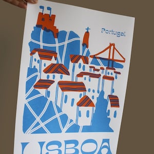 May include: A blue and orange illustrated print of Lisbon, Portugal. The print features a stylized depiction of the city's skyline, including the iconic 25 de Abril Bridge and the Alfama district. The text "LISBOA" is printed in blue at the bottom of the print.