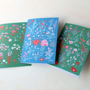 Op de afbeelding: Drie wenskaarten met een bloemenontwerp. Elke kaart heeft een groene achtergrond met witte en roze bloemen en een blauwe achtergrond met witte en rode bloemen. De tekst "Happy Birthday" is in goud op elke kaart gedrukt.