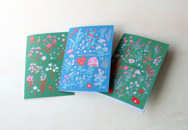 Op de afbeelding: Drie wenskaarten met een bloemenontwerp. Elke kaart heeft een groene achtergrond met witte en roze bloemen en een blauwe achtergrond met witte en rode bloemen. De tekst "Happy Birthday" is in goud op elke kaart gedrukt.