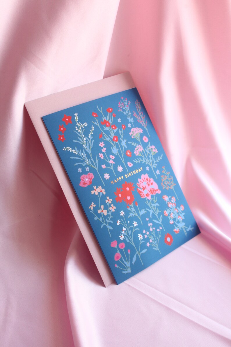 Op de afbeelding: Een blauwe kaart met een bloemenpatroon en de tekst "Happy Birthday" in gouden letters.