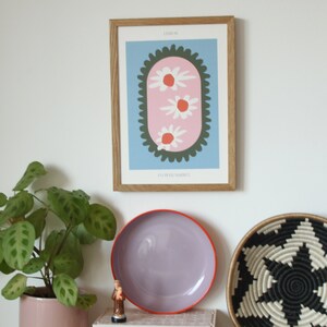 May include: A framed print with a light blue background and a pink oval with three white flowers with orange centers. The oval is surrounded by a dark green scalloped border. The text "LISBON" is at the top of the print and "FLOWER MARKET" is at the bottom.
