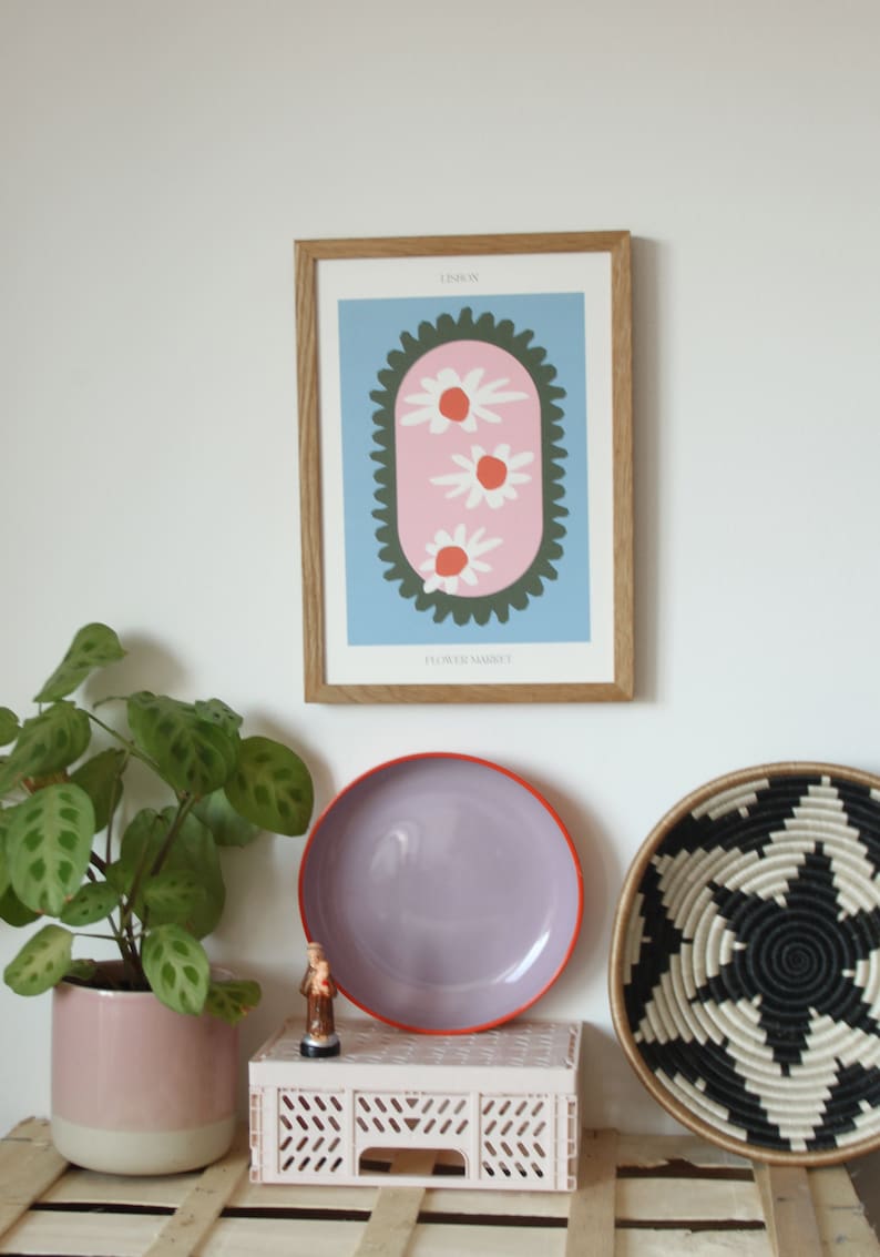 May include: A framed print with a light blue background and a pink oval with three white flowers with orange centers. The oval is surrounded by a dark green scalloped border. The text "LISBON" is at the top of the print and "FLOWER MARKET" is at the bottom.