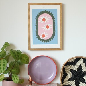 May include: A framed print with a blue background and a pink oval with three white flowers with orange centers. The oval is surrounded by a green border with a scalloped edge. The text "LESLIE" and "FLOWER MARKET" are printed below the image.