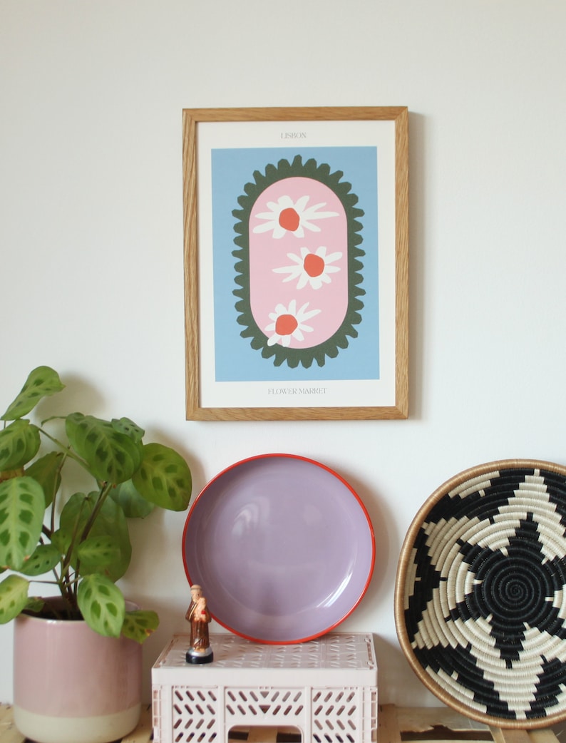 May include: A framed print with a blue background and a pink oval with three white flowers with orange centers. The oval is surrounded by a green border with a scalloped edge. The text "LESLIE" and "FLOWER MARKET" are printed below the image.
