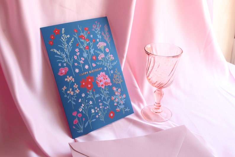 Op de afbeelding: Een blauwe kaart met een bloemenpatroon en de tekst "Happy Birthday" in gouden letters. De kaart ligt op een roze satijnen oppervlak naast een roze glas.