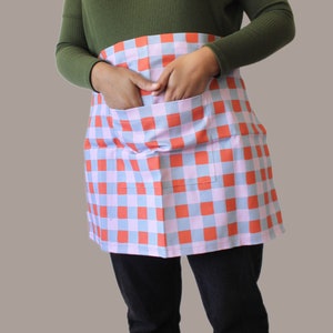 A red, blue and lilac half apron. The apron is made from a cotton linen blend and there is a pocket on the front. Inside the pocket are the model's hands