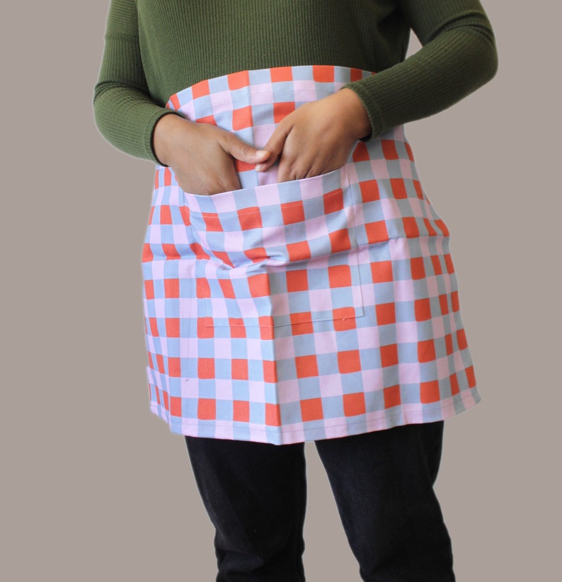 A red, blue and lilac half apron. The apron is made from a cotton linen blend and there is a pocket on the front. Inside the pocket are the model's hands