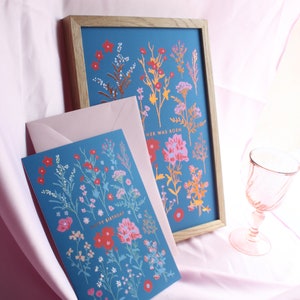 Op de afbeelding: Een blauwe kaart met een bloemenontwerp en de tekst "Happy Birthday" in gouden letters. De kaart ligt op een roze oppervlak naast een ingelijst drukwerk met een vergelijkbaar bloemenontwerp en de tekst "A Flower Was Born".