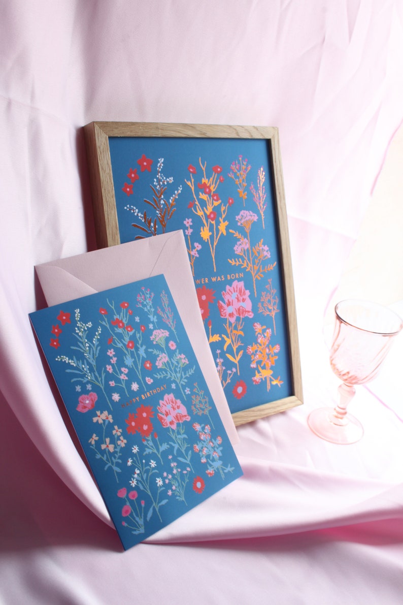 Op de afbeelding: Een blauwe kaart met een bloemenontwerp en de tekst "Happy Birthday" in gouden letters. De kaart ligt op een roze oppervlak naast een ingelijst drukwerk met een vergelijkbaar bloemenontwerp en de tekst "A Flower Was Born".