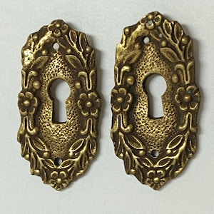 Op de afbeelding: Twee antieke messing sleutelgatdeksels. Elke afdekking heeft een ovale vorm met een sleutelgat in het midden. De afdekkingen zijn versierd met bloemen- en bladerpatronen langs de randen. Het messing heeft een warme, verouderde patina.