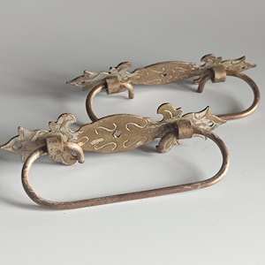 Könnte beinhalten: Zwei Schubladengriffe im antiken Stil mit einem gealterten Gold-Finish. Jeder Griff hat ein verziertes, geschwungenes Design mit einem Bügelgriff. Die dekorative Hardware ist ideal für Schränke, Kommoden oder andere Möbel.