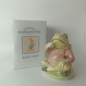 Può includere: Statuetta in ceramica di una rana con cappotto rosa e pantaloni verdi, seduta su una base verde. Dalla collezione Royal Albert "The World of Beatrix Potter", con la scatola visibile.